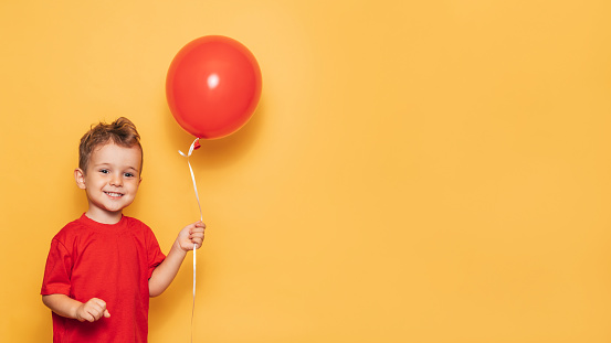 A happy Caucasian boy isolated on a bright yellow background holds a red balloon in his hands. A place for your text or advertisement