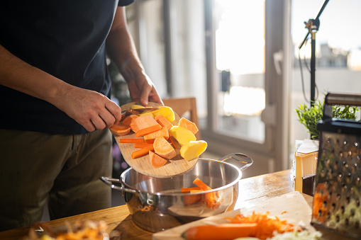 Close-up shot of unrecognizable man moving chopped vegetables from cutting board to pot while preparing lunch at home