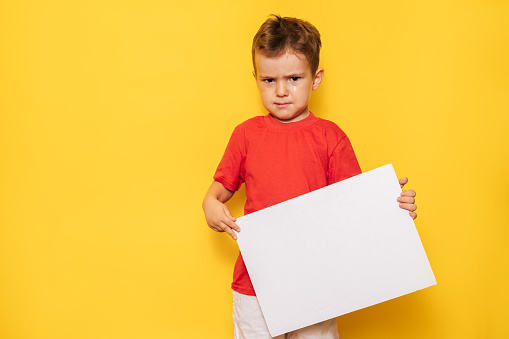 Studio portrait of a disappointed boy with a clean white poster in his hands on a bright yellow background, with a place for your text or advertising.