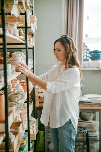 istock Young woman manager of pottery workshop arranges products on shelf 1938972742