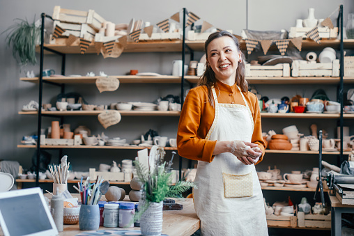 Portrait of a beautiful woman running a pottery studio. Smiling woman potter in apron against background of rack with clay products in workshop