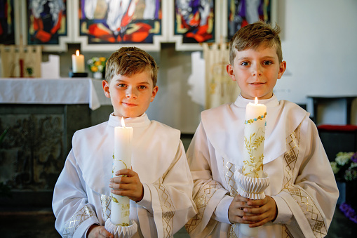 Two little kids boys receiving his first holy communion. Happy children holding Christening candle. Tradition in catholic curch. Kids in a church near altar. Siblings, brothers in white gowns