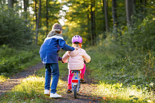 Cute little preschool girl in safety helmet riding bicycle. School kid boy, brother teaching happy healthy sister child cycling and having fun with learning bike. Active siblings family outdoors