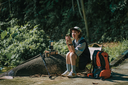 Asian young chinese woman enjoys nature texting with smartphone and boiling coffee resting beside waterfall after hiking trip in weekend morning