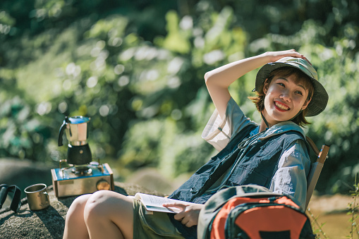 Asian Chinese young woman enjoy reading book with outdoor nature beside river during weekend morning camping