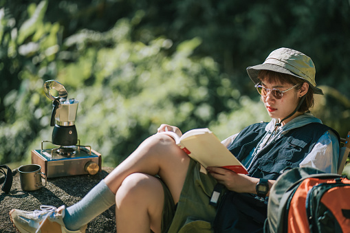 Asian Chinese young woman enjoying me time reading book outdoor nature beside river during weekend morning camping