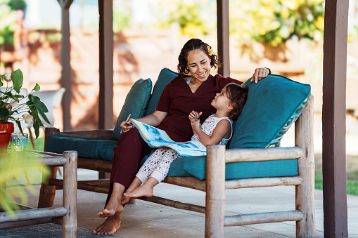 An Eurasian female nurse of Hawaiian descent sits on the lanai at home in Hawaii and helps her cheerful and cute preschool age daughter with schoolwork.