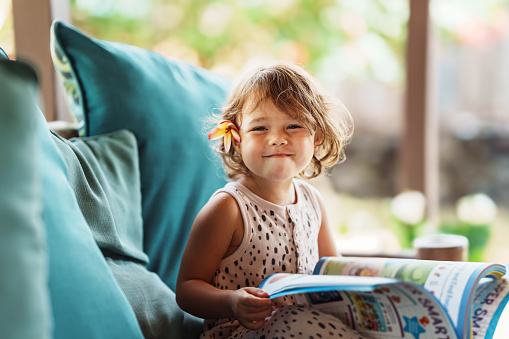 A cute Eurasian three year old girl of Hawaiian and Chinese descent sits on the sofa on the lanai of her home in Hawaii and smiles directly at the camera while holding her school workbook on her lap.