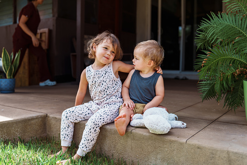 An adorable three year old Eurasian girl lovingly embraces her one year old brother while sitting together outside on the back patio of their home in Hawaii.