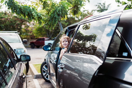 Portrait of a cute three year old Eurasian girl smiling directly at the camera while climbing out of the open passenger door of her family's mini van which is parked in the parking lot of a shopping center in Hawaii.