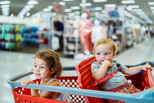 A cute one year old boy and three year old girl of Eurasian descent sit in a shopping cart while shopping with their parents in the clothing section of a superstore.