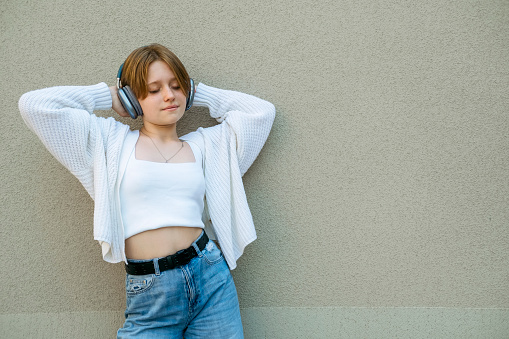 Portrait of a teenage girl in headphones against a gray wall. A teenager in jeans and a white jacket. Without an emotional face, indifference