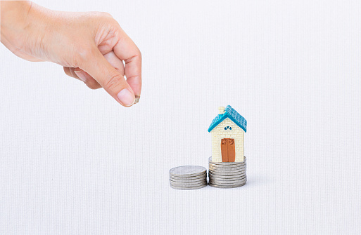 Miniature house on coin stack with hand holding coin on white texture background, property investment, real estate business, buy new house