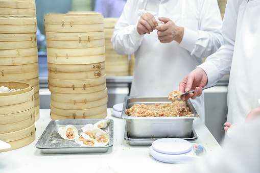 A group of chefs are making soup dumplings and siomai in a clean and bright kitchen. Soup dumplings and siomai are famous snacks in Chinese society. They use stick-shaped flour buns filled with minced pork or shrimp. The chefs wear white aprons, masks and hats and they attach great importance to food hygiene