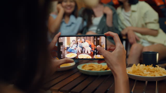 Closeup group of Asian female friends take picture with phone while camping in living room at home in night time. Happy women laughing while talking in front of a tent enjoy leisure activity.