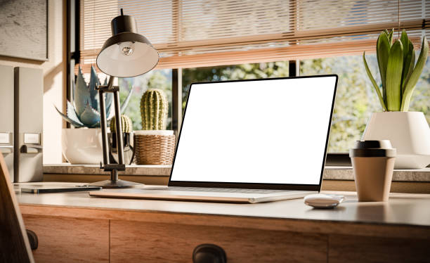 A laptop with a blank screen sits on a stylish wooden desk within a loft-style interior, with green spaces in the background visible through the window