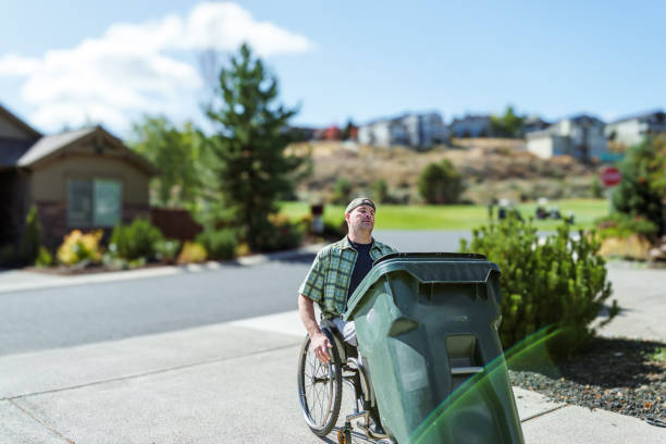 An adult Caucasian man with a physical disability and using a wheelchair takes the garbage can out to the side of a residential street in Oregon.