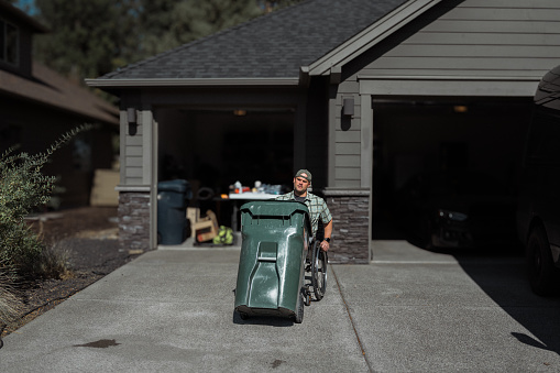 A middle aged man with a disability who uses a wheelchair brings the garbage can down his driveway to the street for pickup on a beautiful sunny day in Central Oregon.