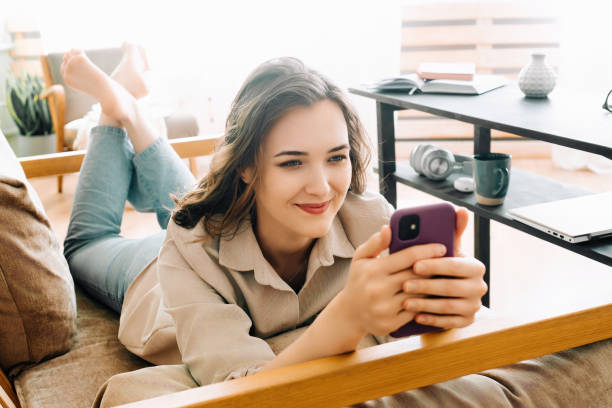 cheerful millennial woman relaxing on sofa, glancing at phone, reading messages, chatting, and enjoying free time. cozy leisure at home with happy young woman engaging in digital communication. - news of the world imagens e fotografias de stock