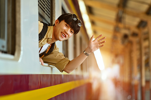 Happy young Asian man waving hand to friend through train window before leaving.