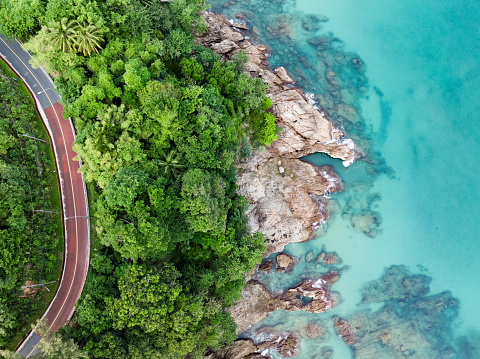 View from above, stunning aerial view of a road that runs alongside a rocky coastline surrounded by palm trees and bathed by a turquoise water. Phuket, Thailand.