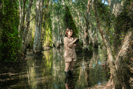 Asian ecologist standing in the flood area and exploring the rainforest to collect data for research while writing the detail on her notebook. Active female dedicating her life on conserving the environment.