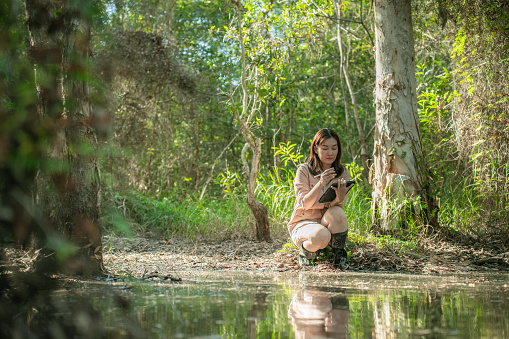 Asian female environmental researcher sitting near the swamp and collecting the data by writing on her notebook for her dissertation about conserving the environment in the rainforest.