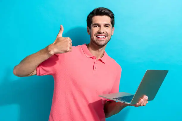 Portrait of cheerful young guy in pink t shirt shows thumb up like symbol holding laptop xiaomi isolated on aquamarine color background.