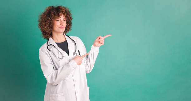 Cheerful woman doctor pointing at blank green wall stock photo
