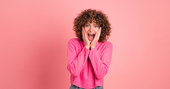 Astonished young curly haired female in makeup and opened mouth looking at camera while standing with folded hands and touching hair on pink background
