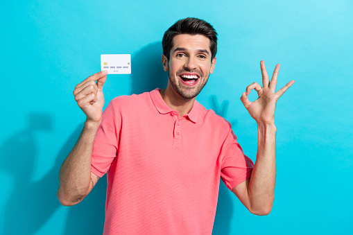 Portrait of young funny person guy in pink t shirt shows okey sign recommend debit card advantages isolated on aquamarine color background.