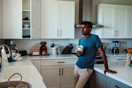 Portrait of a handsome Indian man looking out the kitchen window with a contemplative expression while holding a cup of water.