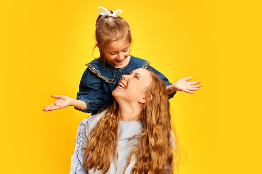Little girl, daughter surprising her mother against yellow studio background. Celebrating women's holiday. Concept of Mother's Day, International Happiness Day, motherhood, childhood