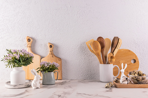 Wooden kitchen utensils made of eco-friendly materials, spring flowers in a vase and jug, Easter bunny figurines on a marble countertop. Front view