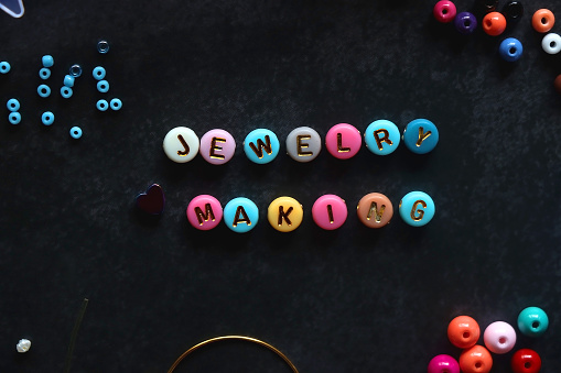 Colorful beads and various jewelry making supplies on dark background. Letter beads spelling JEWELRY MAKING. Top view.
