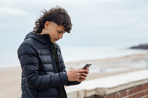 A portrait of a teenager wearing casual clothing on an overcast summer day in Whitley Bay, Northeastern England. They are standing and looking concerned as they use their smartphone.
