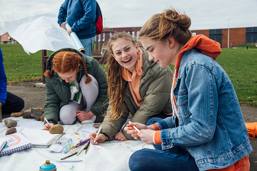 A group of teenage friends wearing casual clothing on an overcast summer day in Whitley Bay, Northeastern England. They are gathered on the promenade by the sea where they use different arts and crafts products to create sustainability artwork to spread a message of environmental conservation for Earth Day.