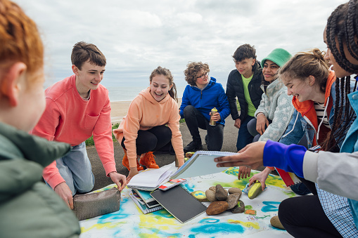 A group of teenage friends wearing casual clothing on an overcast summer day in Whitley Bay, Northeastern England. They are gathered on the promenade by the sea where they use different arts and crafts products to create sustainability artwork to spread a message of environmental conservation for Earth Day.