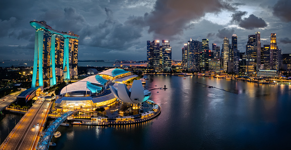 Singapore Urban Cityscape - Skyline at Marina Bay in colorful twilight after sunset. Drone Point of view panorama. Green illuminated Towers. Stitched Panorama DJI Mavic 3 Pro. View over the glowing Singapore Waterfront Modern Skyscrapers and light reflexions under moody twilight skyscape. Singapore, Southeast Asia, Asia