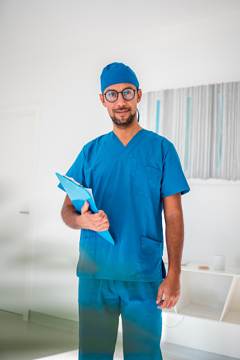 Professional Hispanic male surgeon in blue surgical attire holding a clipboard with a confident smile, standing in a bright medical office setting.
