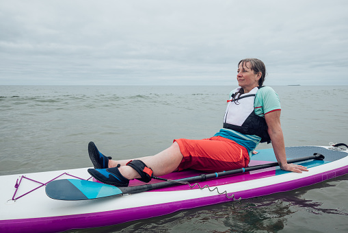 Mature woman paddle boarding in the sea at the coast Alnmouth, Northumberland. The sky is overcast and she is having fun in the waves, sitting reclined on the board.