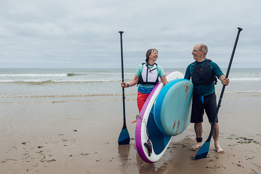 Mature couple paddle boarding at the coast Alnmouth, Northumberland. They are on the beach walking side by side carrying their boards under their arms. They are laughing while looking at each other.