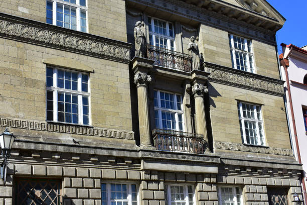 Facade of the historic building is made of grey stone with decorative elements on the facade, statues, columns and windows in white frames. Ancient architecture. Poland, Torun, August 2023