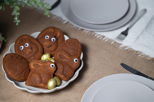 Close-up of Easter muffins shaped as bunnies and eggs with eyes, chocolate eggs, framed by green plant. View from above on table setting with empty gray plates and cutlery