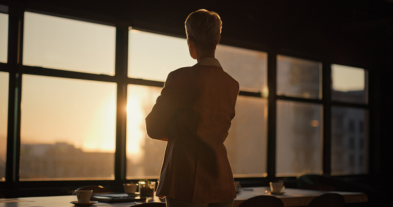 Sunset, window and back of woman in the office thinking, planning and brainstorming for project. Decision, choice and professional female person with memory or reflection at workplace in evening.