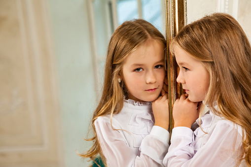 Face of child cover girl model 6-7 year old looking at mirror, thought look at camera. Portrait of cute kid girl in white attire pose in room. Youth generation z emotion concept. Copy ad text space