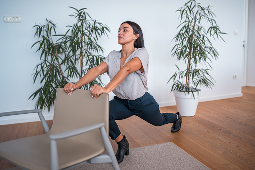 A Hispanic female employee in a business casual outfit performs a leg stretch using a chair at the workplace, promoting fitness and health at work.