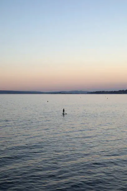 A female lone paddleboarder is seen almost in silhouette on the calm summer sea at sunset. This was taken in June, Summer 2020 when being alone and getting out and being active was very important. The Purbeck hills are in the background.