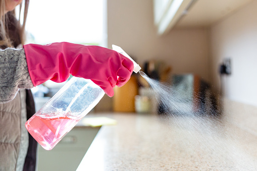 Close up image depicting a woman's hand - wearing a protective pink rubber glove - spraying surface cleaner onto the kitchen countertop.