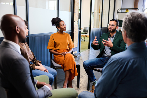 Businessman explaining a concept while talking with a team of diverse businesspeople sitting in a circle during a casual office meeting